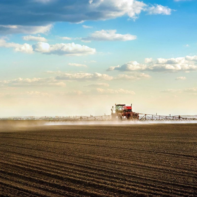 Traktor mit Spr&uuml;harm f&auml;hrt bei sch&ouml;nem Wetter &uuml;ber braunes Feld und spr&uuml;ht.
