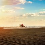 Traktor mit Sprüharm fährt bei schönem Wetter über braunes Feld und sprüht.