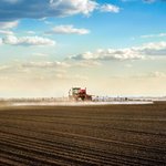 Traktor mit Sprüharm fährt bei schönem Wetter über braunes Feld und sprüht.