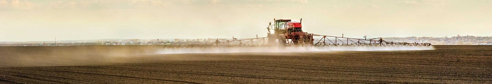 Traktor mit Spr&uuml;harm f&auml;hrt bei sch&ouml;nem Wetter &uuml;ber braunes Feld und spr&uuml;ht.