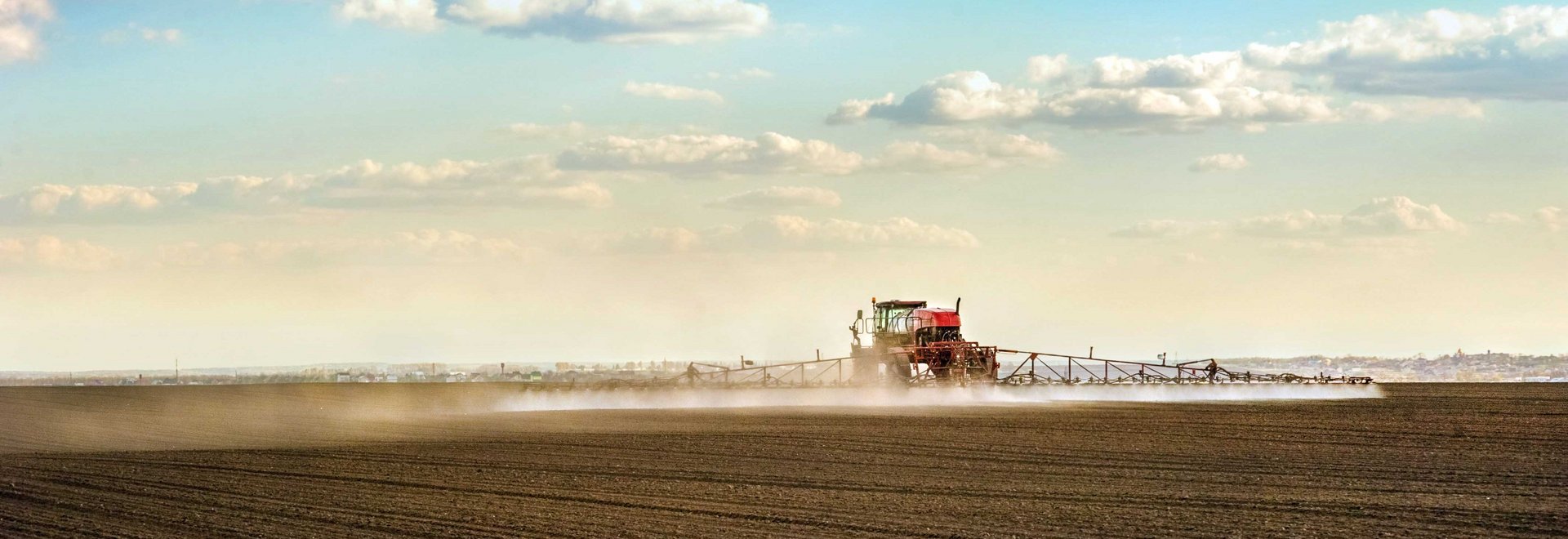 Traktor mit Spr&uuml;harm f&auml;hrt bei sch&ouml;nem Wetter &uuml;ber braunes Feld und spr&uuml;ht.